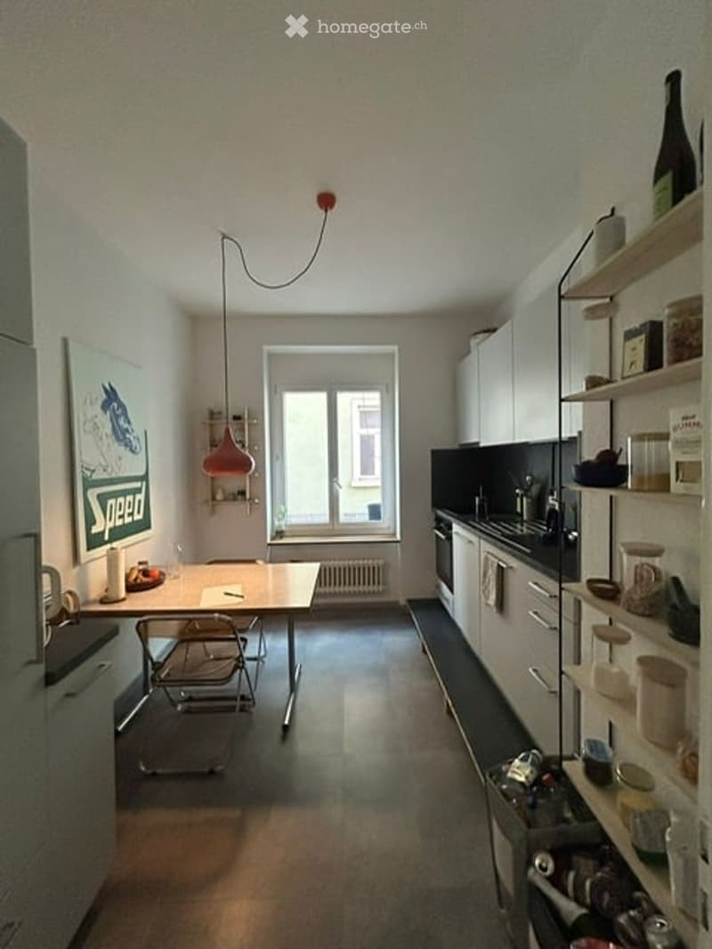 Modern kitchen with white cabinets, black countertops, a central dining table with chairs, and various kitchen items on shelves and counters.
