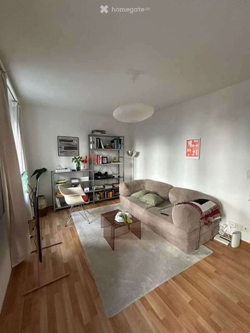 Modern living room with light wooden flooring, a grey sofa, a coffee table, a bookshelf, a white chair, a floor lamp, and a small plant. The room features large windows with white curtains and a round ceiling light.