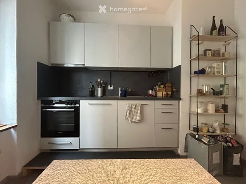 Modern kitchen with white cabinets, black countertops, built-in oven, dishwasher, and a metal shelf with various kitchen items.