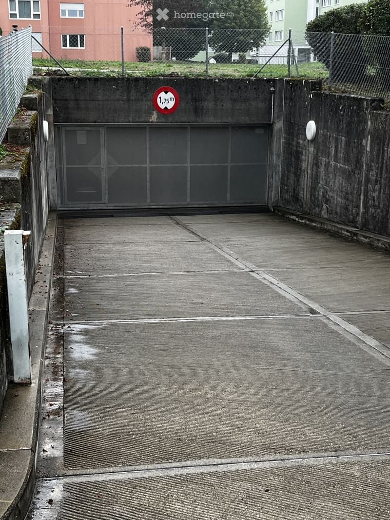 Concrete garage entrance with a height restriction sign of 1.75 m, surrounded by a chain-link fence and adjacent buildings.