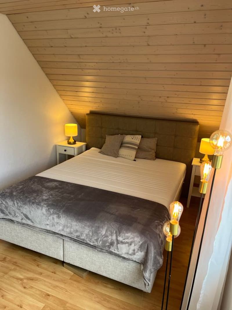 Cozy bedroom with wooden walls and floor, featuring a large bed with a grey duvet and decorative pillows, nightstands with lamps, and warm lighting.