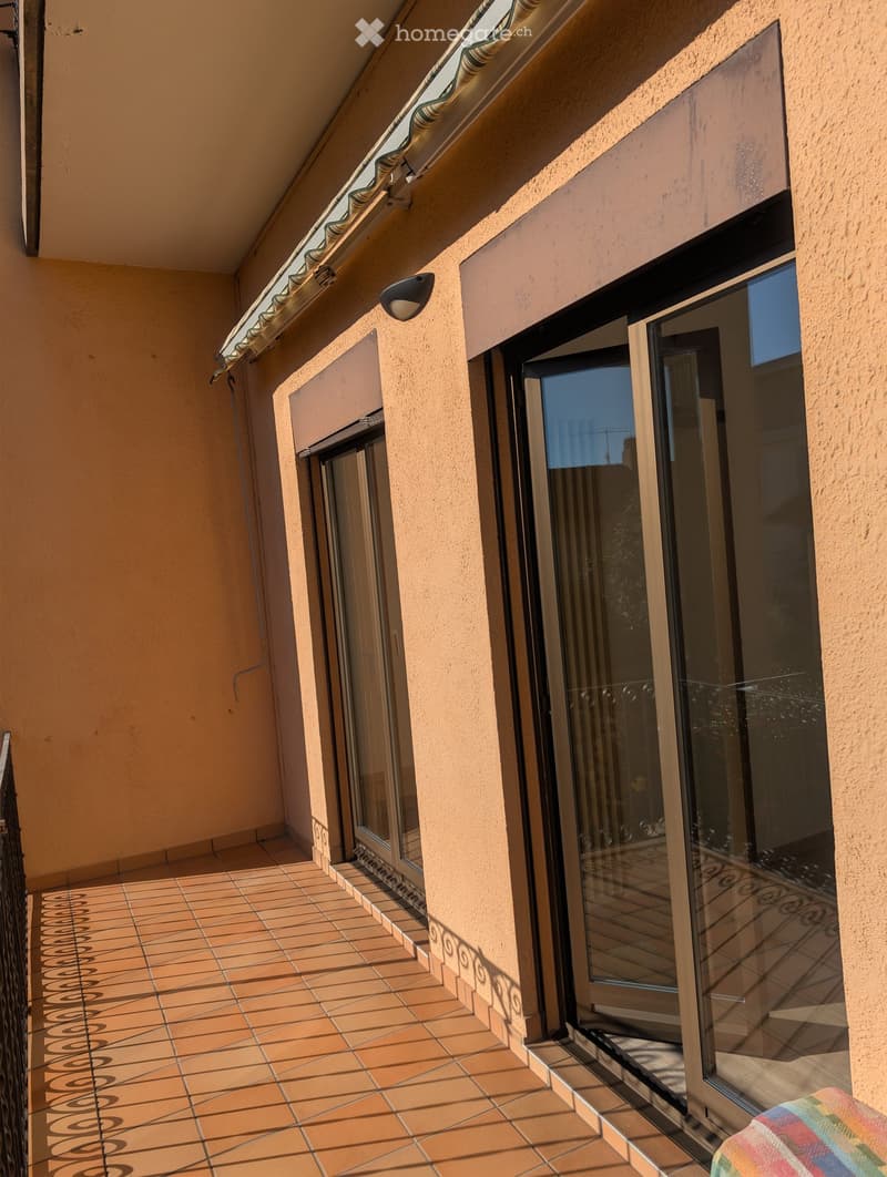 Balcony with tiled floor, glass sliding doors, beige walls, and a small awning above.