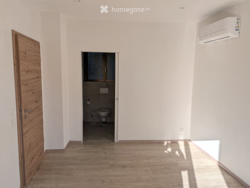 Empty room with light wooden flooring, white walls, and a wooden door leading to a small bathroom with a toilet and sink.