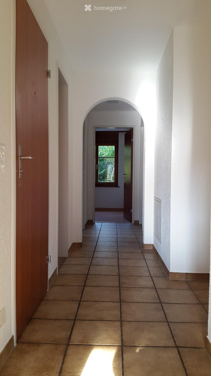 A long, narrow hallway with tiled flooring and white walls, featuring a wooden door on the left and an arched passageway leading to another room with a window showing greenery outside.