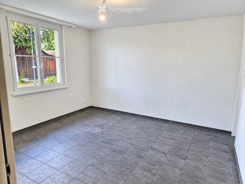 Empty room with grey tiled floor, white walls, and a large window with a view of a wooden fence and greenery outside.