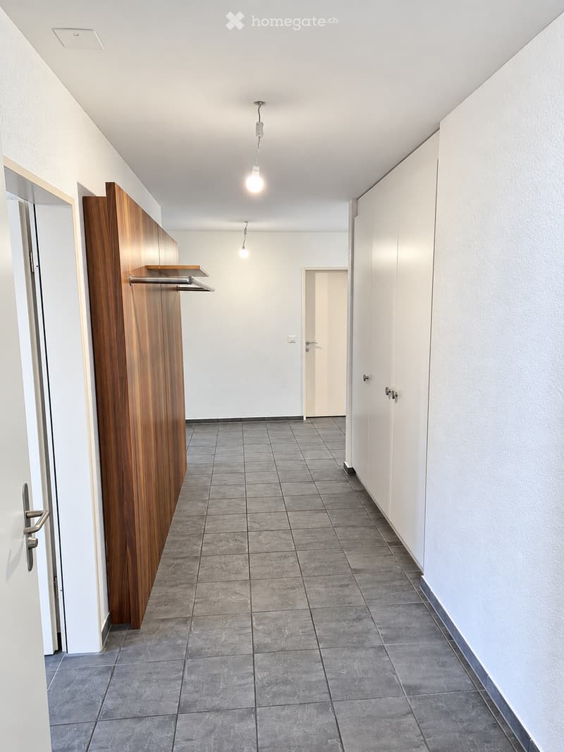 Empty hallway with grey tiled flooring, white walls, and a wooden coat rack on the left side. Two doors are visible, one on each side of the hallway.