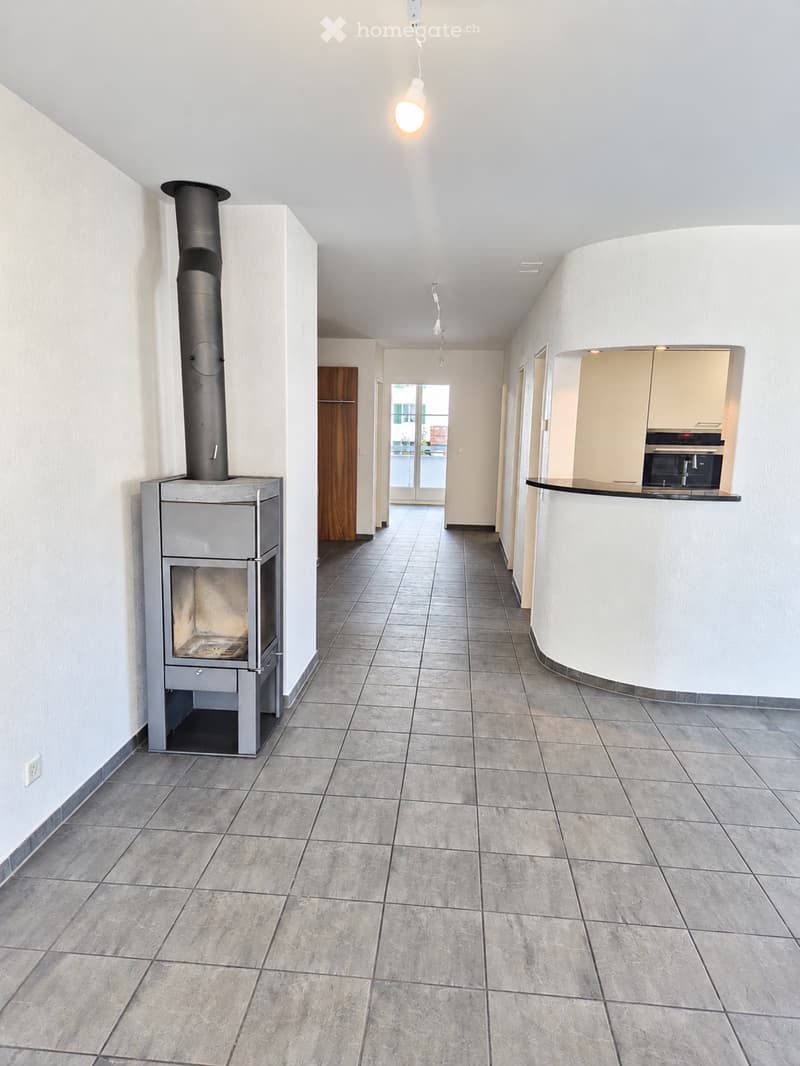 Empty hallway with tiled floor, white walls, and a visible fireplace on the left side. The hallway leads to a kitchen area with cabinets and a countertop on the right.