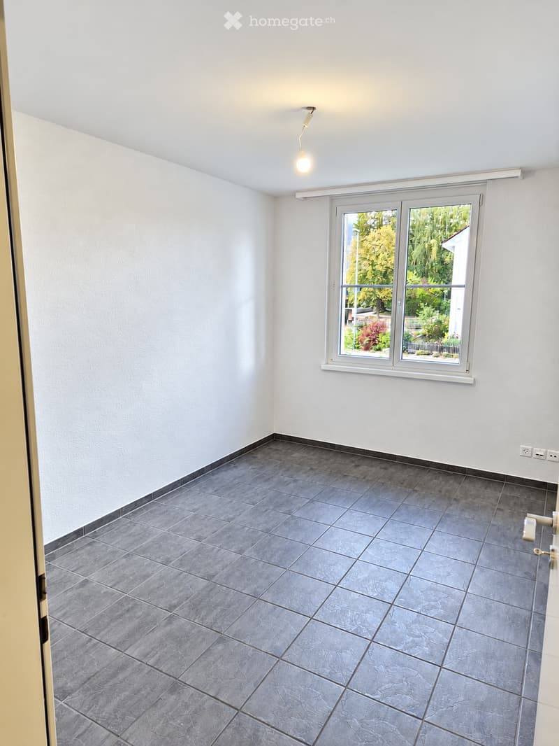 Empty room with grey tiled floor, white walls, and a large window showing greenery outside. No furniture or equipment present.