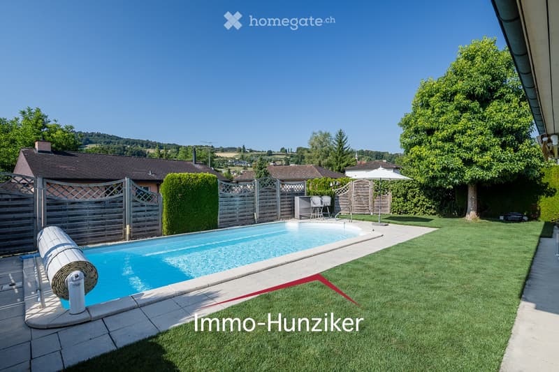Well-maintained garden with a swimming pool, surrounded by a wooden fence, lush greenery, and a clear blue sky.