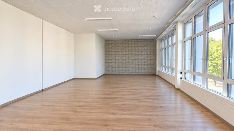 Empty room with light wooden flooring, white walls, large windows, and a brick accent wall.