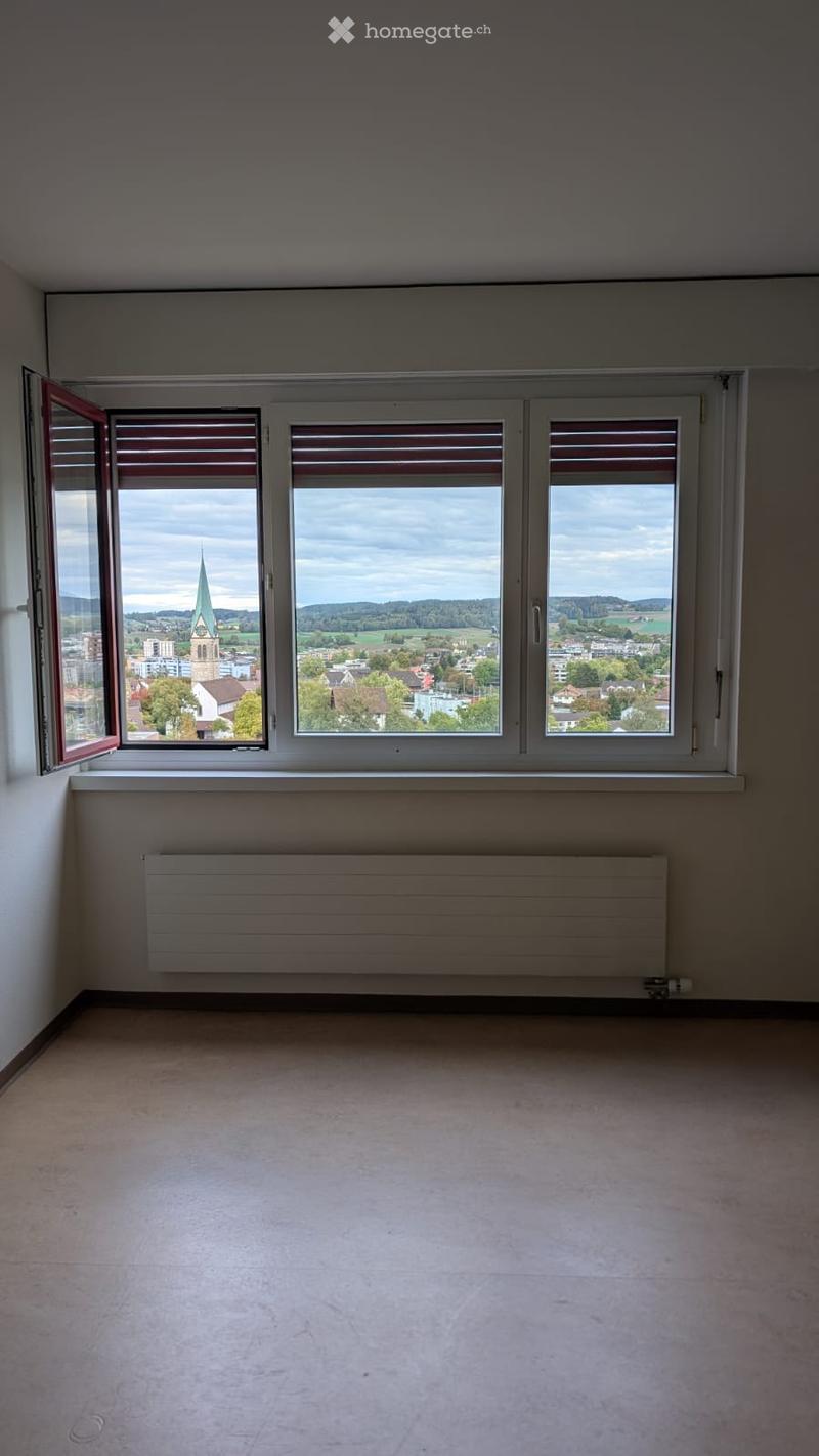 Empty room with large windows offering a scenic view of a town and countryside, featuring a radiator below the windows and a smooth concrete floor.