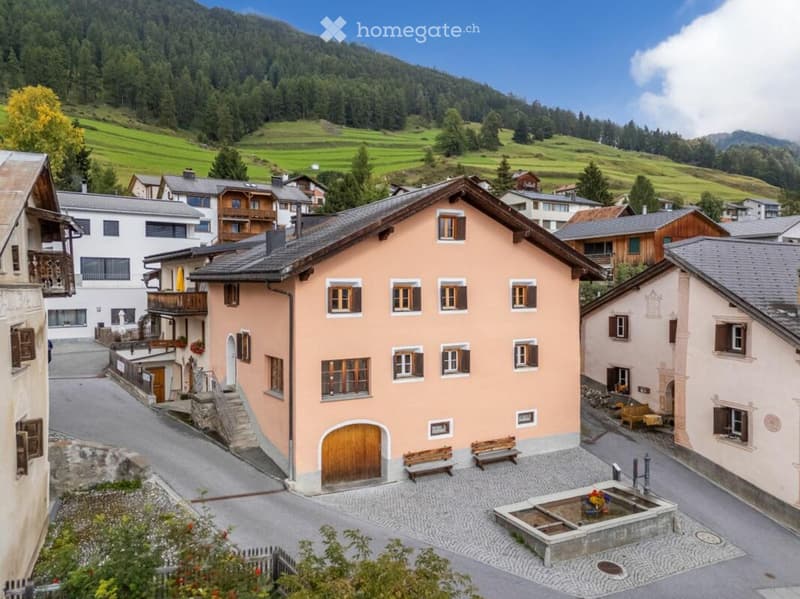 A picturesque village scene with traditional pink houses, wooden balconies, and a central fountain surrounded by benches.