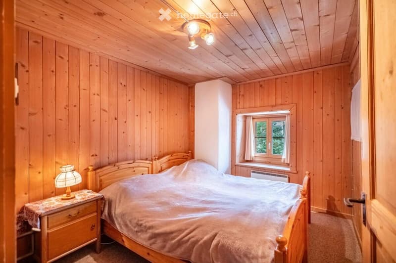 Cozy wooden bedroom with a large bed, nightstand, and lamp. The room features wooden walls and ceiling, a window with curtains, and warm lighting.