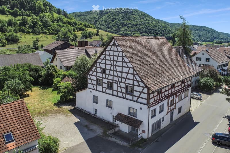 A traditional half-timbered house with a tiled roof, surrounded by other houses and greenery in a rural setting.