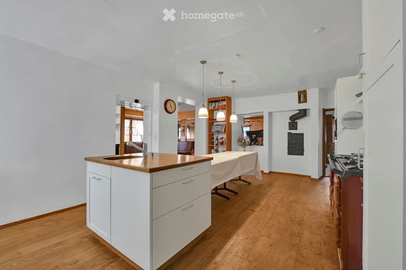Bright kitchen with white cabinets, wooden countertop, and hardwood flooring. Features a sink, faucet, table, fan, oven, bookshelf, and clock.