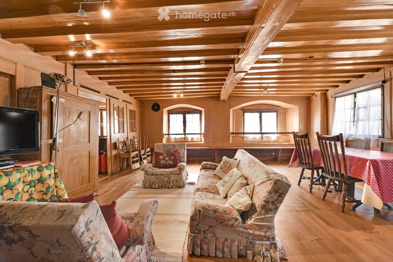 Cozy living room with wooden beams, exposed wooden ceiling, large windows, floral-patterned sofa and armchair, dining table with chairs, television, and floor lamp.