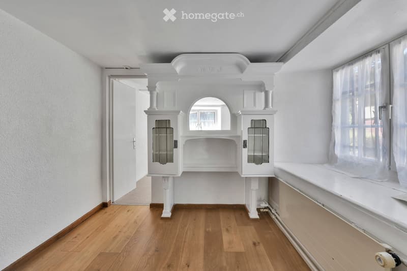 Empty room with wooden flooring, white walls, built-in white cabinetry with glass doors, and a large window with white curtains.
