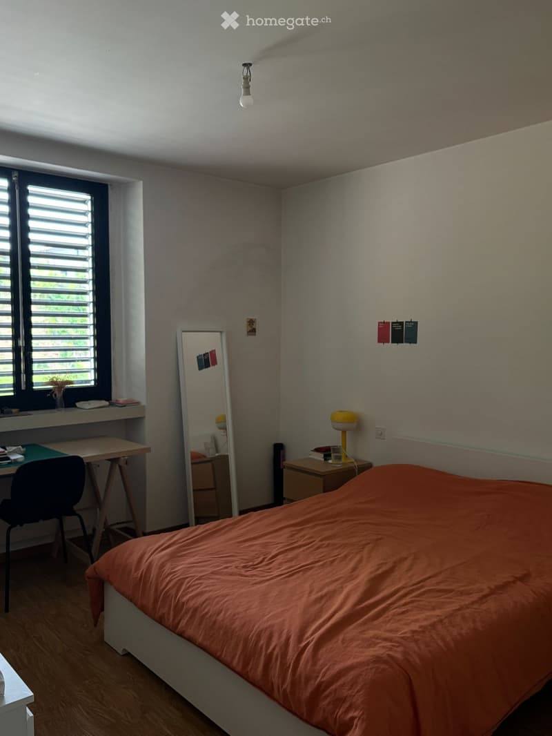 A modern bedroom with a single bed, desk, chair, and nightstand. The room features a large window with blinds, a mirror, and a lamp on the nightstand.