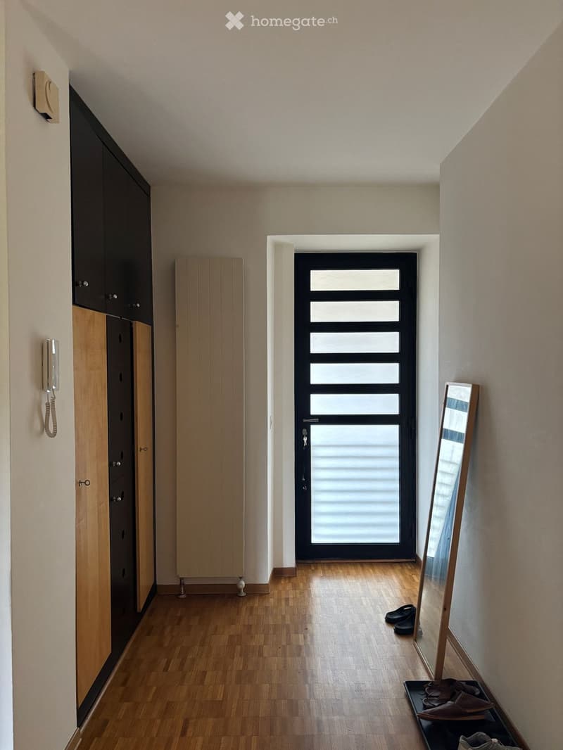 Modern hallway with wooden flooring, built-in wooden and black cabinets, a black door with frosted glass panels, a wall-mounted telephone, a mirror on a stand, and a small shoe rack with shoes and slippers.