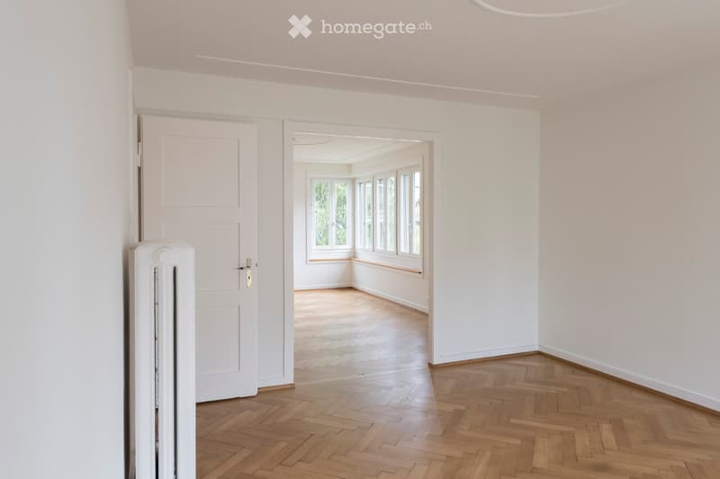 Empty hallway with wooden flooring, white walls, and a door leading to another room with large windows and wooden flooring.