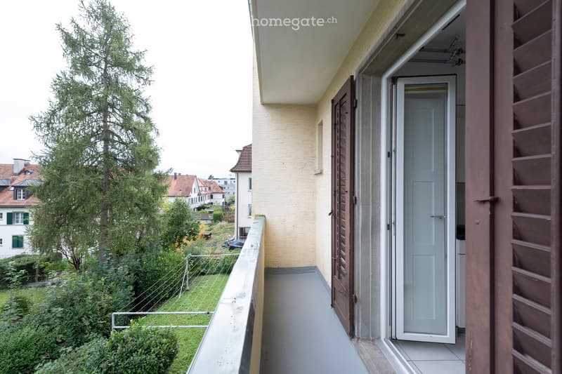 Balcony with open sliding door, overlooking a green garden and neighboring houses.