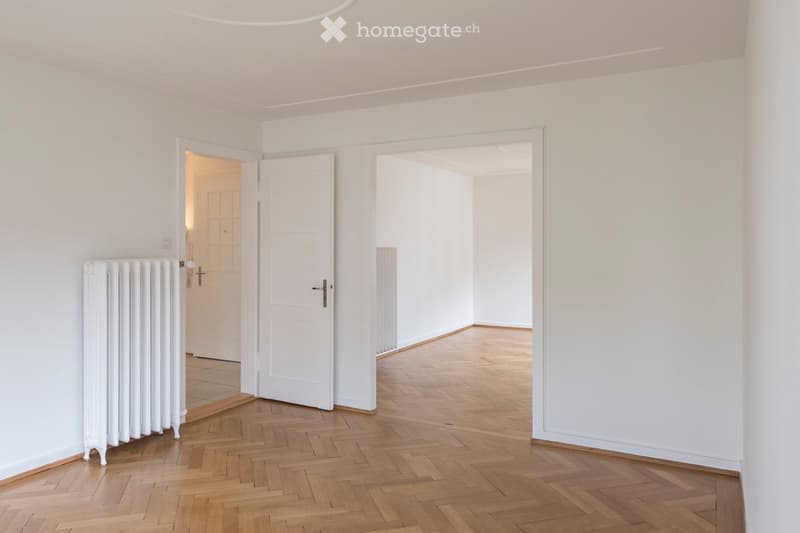 Bright, minimalist hallway with white walls, wooden herringbone flooring, and two white doors leading to adjacent rooms.