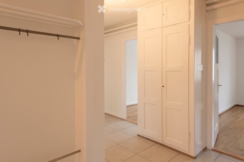 Empty hallway with white walls, tiled floor, and wooden flooring in adjacent room. Features built-in closet with sliding doors and a visible closet rod.