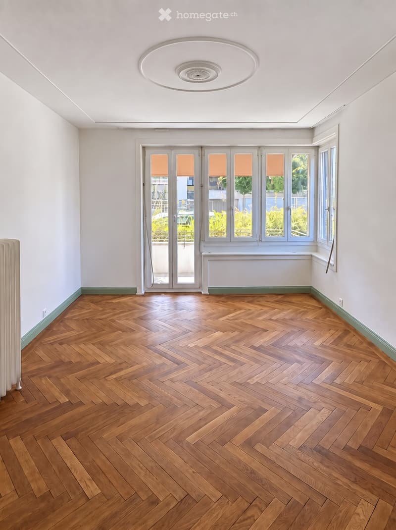 Empty room with herringbone wooden flooring, white walls, large windows, and a sliding glass door leading outside.
