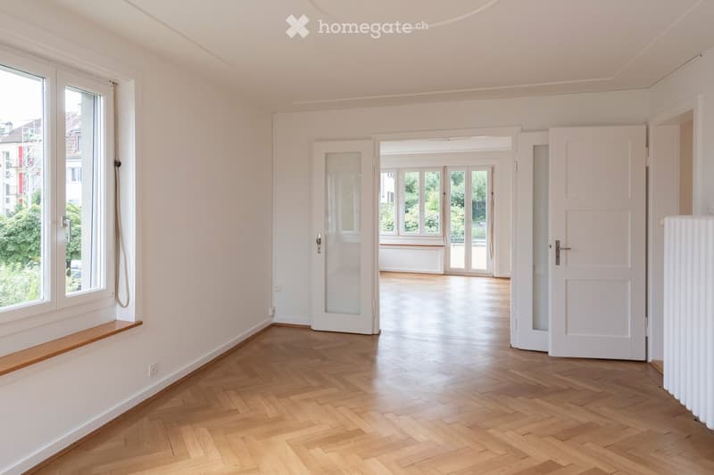 Empty room with wooden flooring, white walls, large windows, and open doors leading to another space.