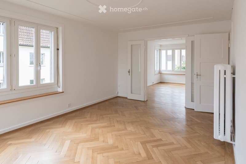 Empty room with light wooden herringbone flooring, white walls, large windows, and a radiator. No furniture or personal items are present.