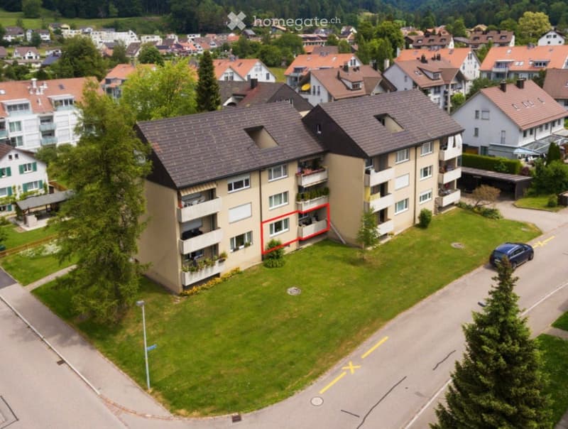 Aerial view of a multi-story residential building with balconies, surrounded by greenery and neighboring houses.