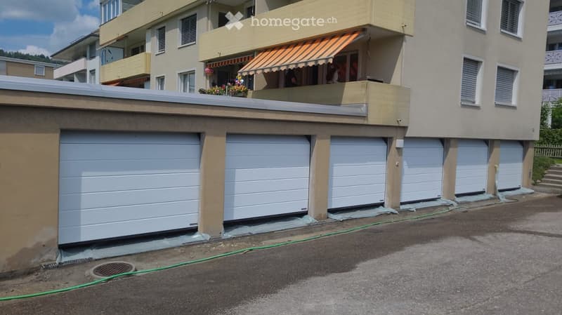 Multi-story residential building with a large garage area featuring multiple garage doors, a small balcony with an awning, and flower boxes.