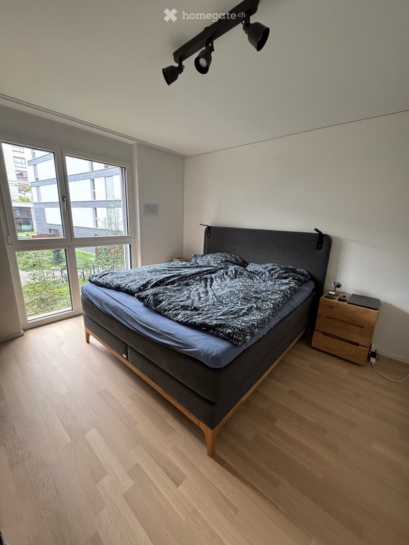 Modern bedroom with a large bed, nightstand, laptop, and a sleek wall-mounted lamp. The room features light wooden flooring and large windows offering a view of greenery.