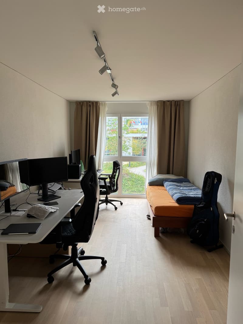 A furnished room with a desk, ergonomic chair, two screens, a bed with a mattress, and a backpack. The room has wooden flooring and large windows with curtains.