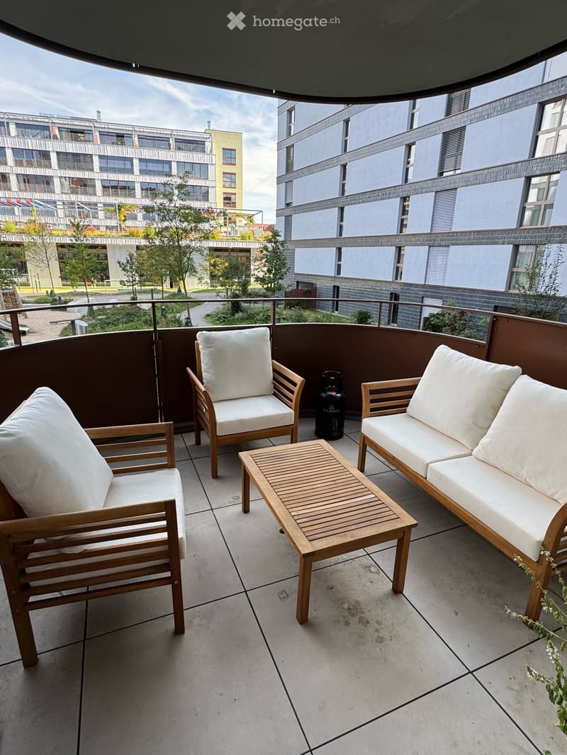Balcony with wooden furniture including a sofa, chairs, and a small table, surrounded by modern buildings and greenery.