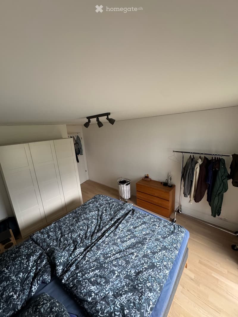 A tidy bedroom with a bed covered in a blue patterned duvet, a white wardrobe, a wooden dresser, a clothing rack with hanging garments, and a small trash bin near the dresser.