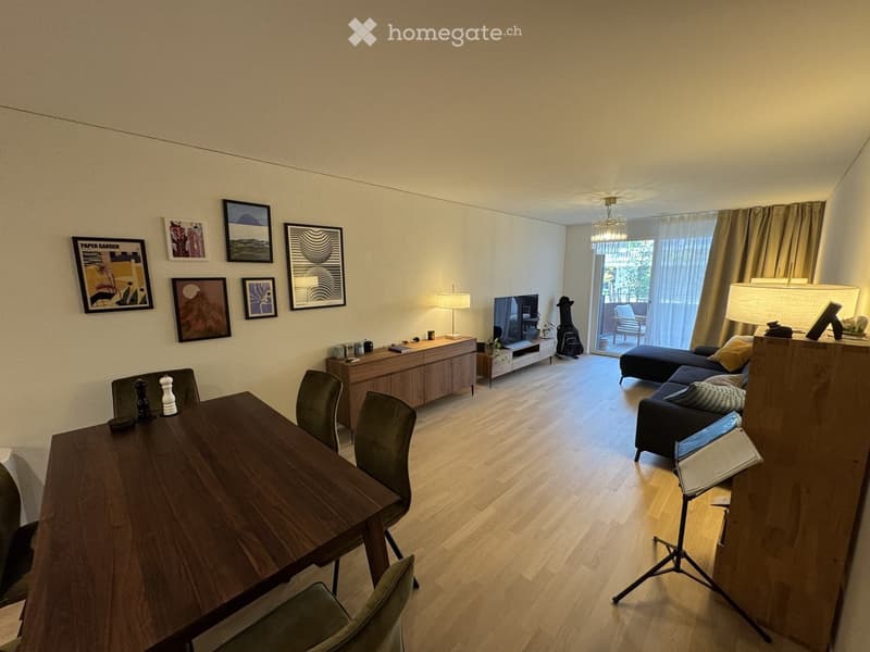 A modern living room with a wooden floor, a sofa, a dining table with chairs, a TV, and several lamps. There are picture frames on the wall and a large window with curtains leading to a balcony.