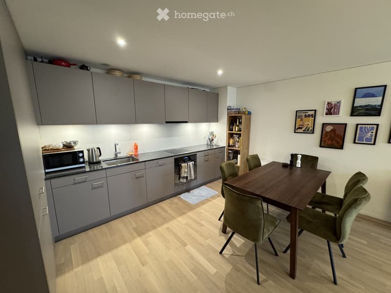 Modern kitchen with grey cabinets, wooden floor, microwave, kettle, sink, dishwasher, dining table with four chairs, and framed pictures on the wall.