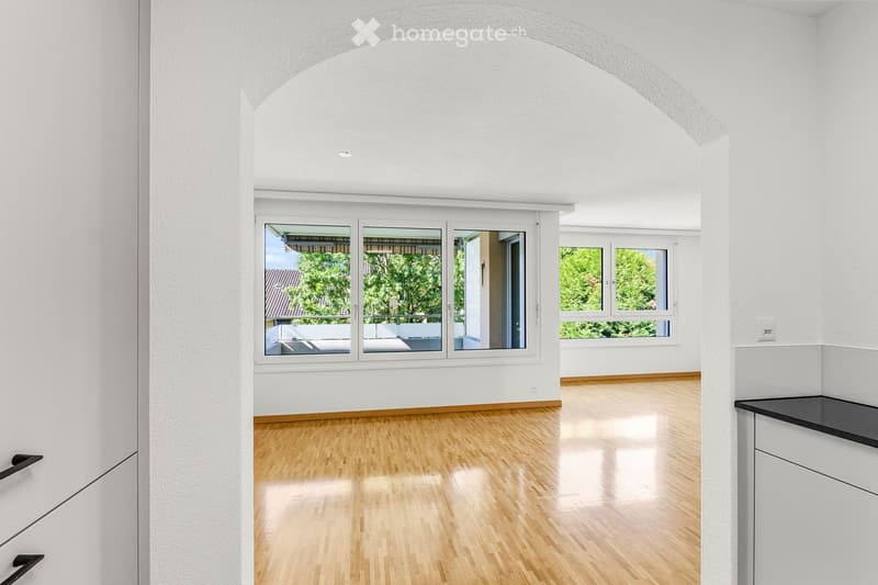Bright, empty room with wooden flooring, white walls, large windows, and a balcony view of greenery.