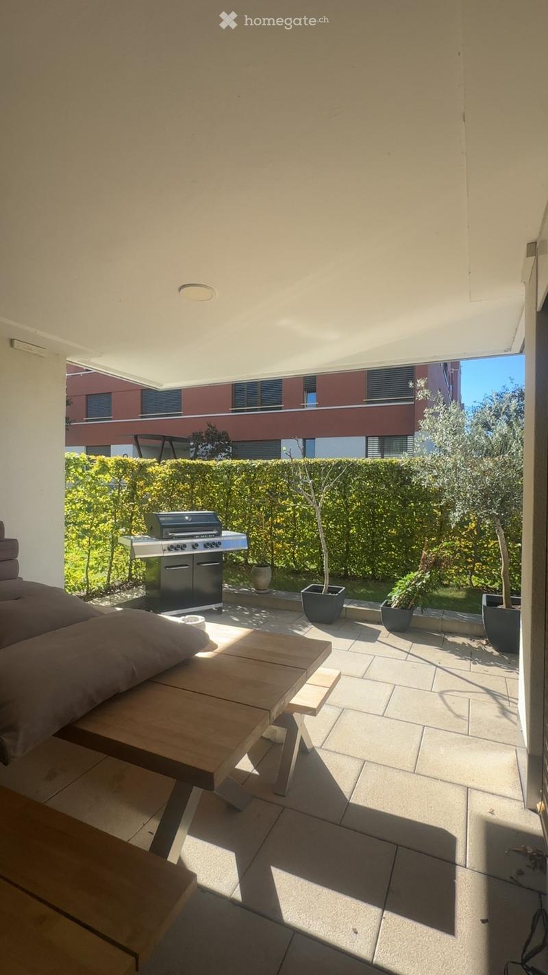 Balcony with a wooden table, sofa, grill, and several potted plants, overlooking a landscaped garden area.