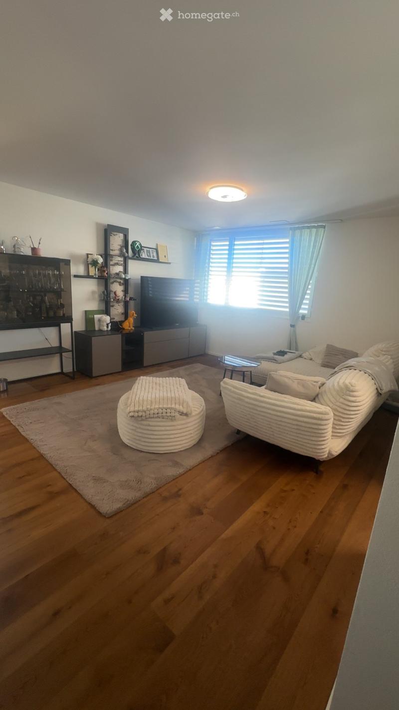 Modern living room with wooden flooring, large window with curtains, sofa, coffee table, ottoman, TV, cabinet, and shelves.