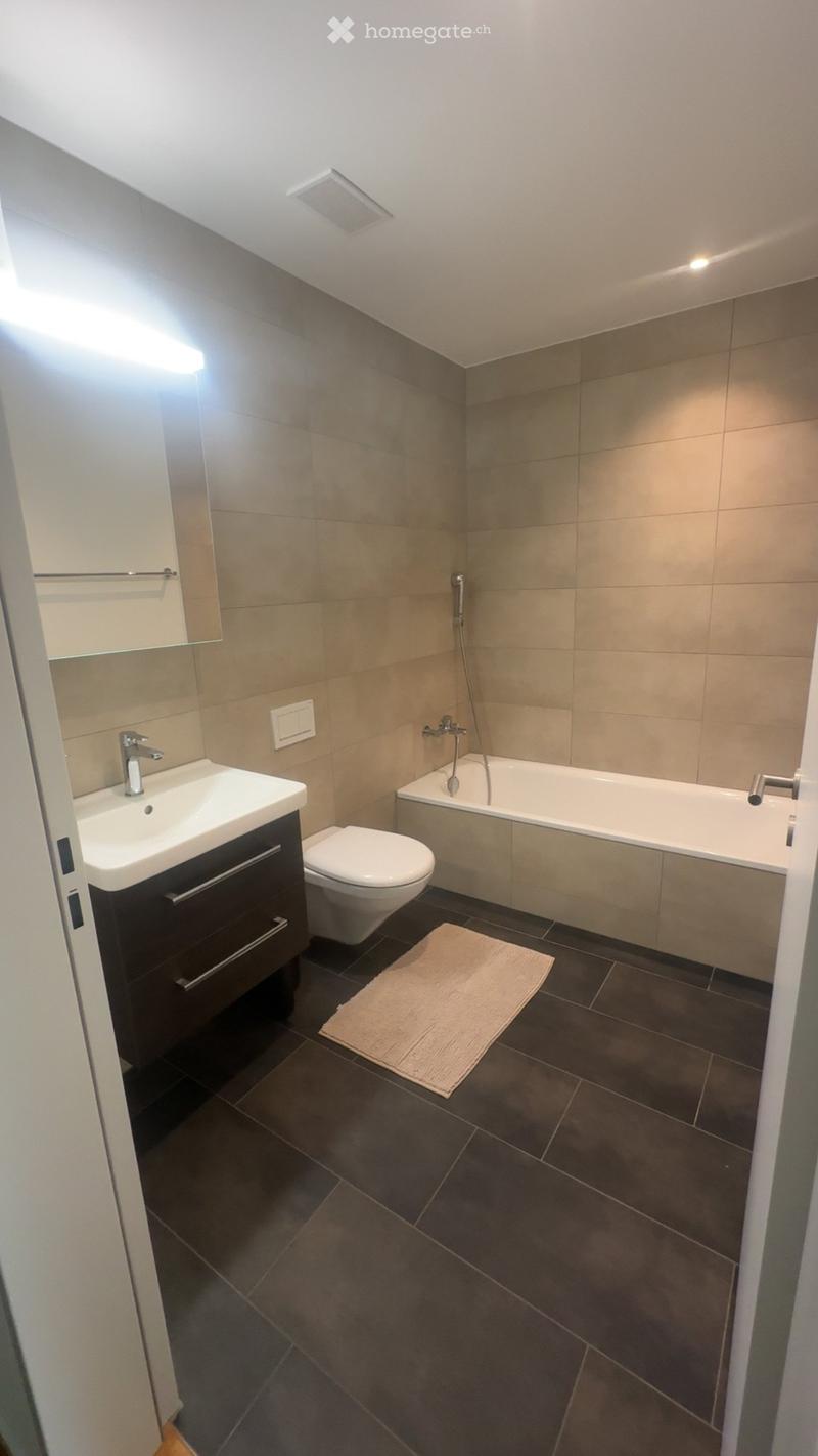 Modern bathroom with dark tiled floor, beige tiled walls, a white sink with a dark cabinet, a toilet, a bathtub with a showerhead, a mirror, and a small beige rug.
