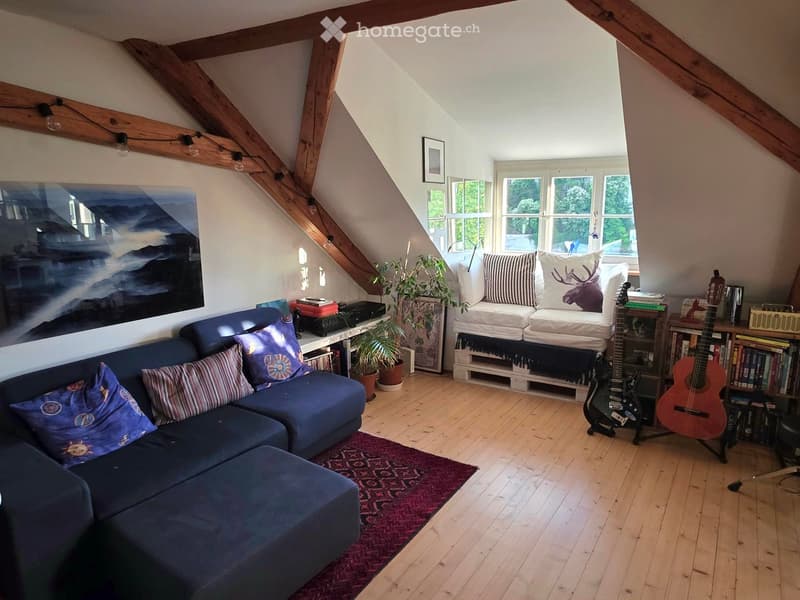 Furnished Room. Cozy attic living space with exposed wooden beams, large windows, blue sectional sofa, ottoman, guitars, bookshelf, and potted plants.