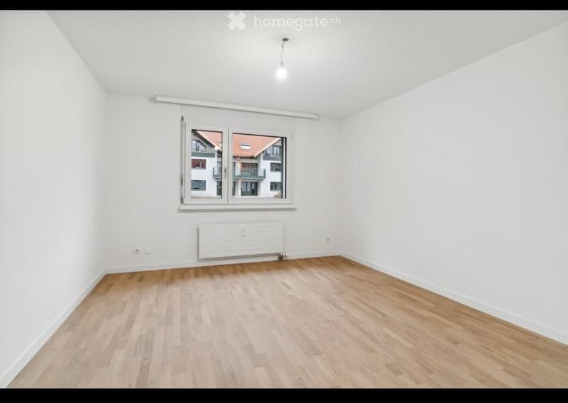 Empty room with white walls, wooden flooring, and a large window showing a view of neighboring buildings.