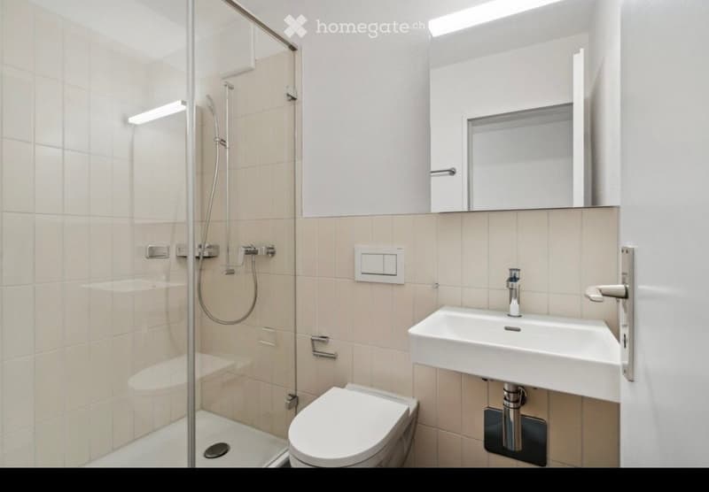 Modern bathroom with a white toilet, sink, and glass-enclosed shower. Features beige tiles, a large mirror, and sleek fixtures.