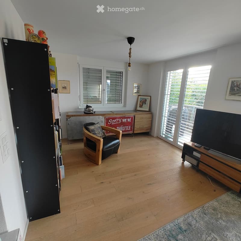 Bright living room with wooden flooring, large windows, a black leather armchair, a TV on a wooden stand, and a bookshelf with books and decorative items.