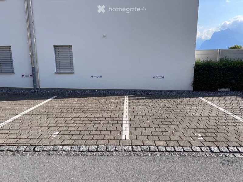 A modern building exterior with a paved parking area, white walls, and two windows with blinds. The parking area is marked with white lines and has a clean, organized appearance.