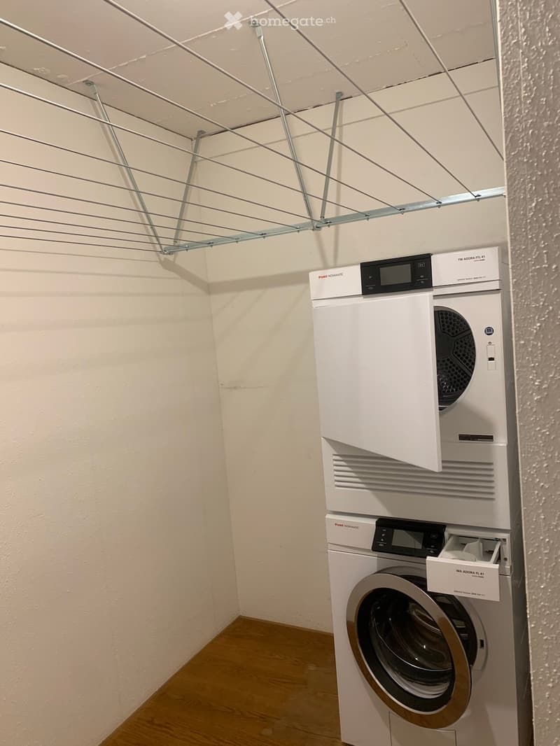 A small, clean bathroom with a stacked washing machine and dryer unit, white walls, and a wooden floor.