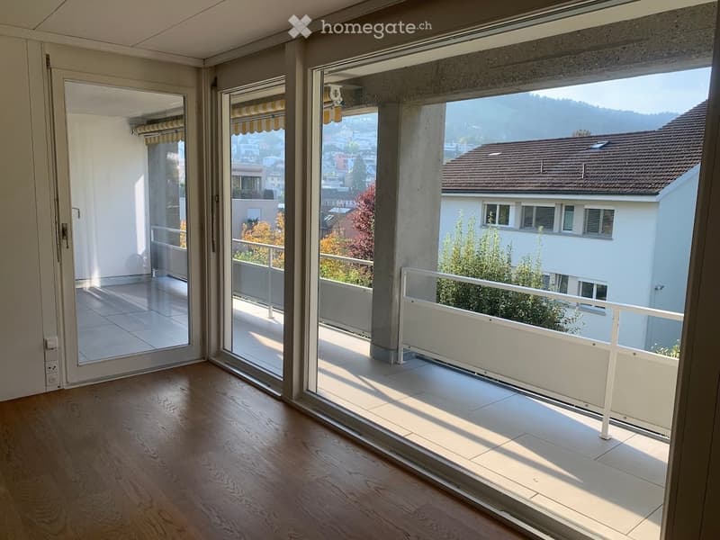 Bright, unfurnished room with large glass doors leading to a balcony, offering a scenic view of the city and surrounding hills.