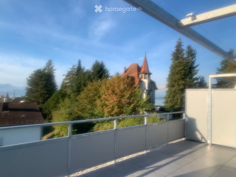 Balcony with tiled floor, metal railing, and scenic view of trees, a red-roofed building, and distant mountains.